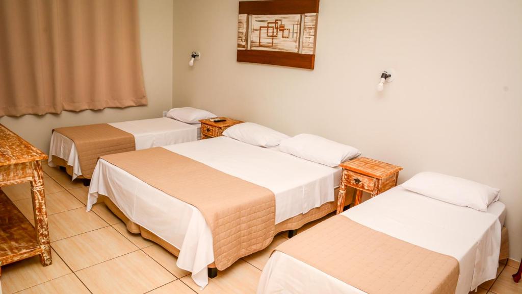 Um quarto no Hotel Winner com uma cama de casal e duas camas de solteiro, cada uma com lençóis brancos e coberturas bege, e mesas de cabeceira de madeira, para representar hotéis em Barretos.