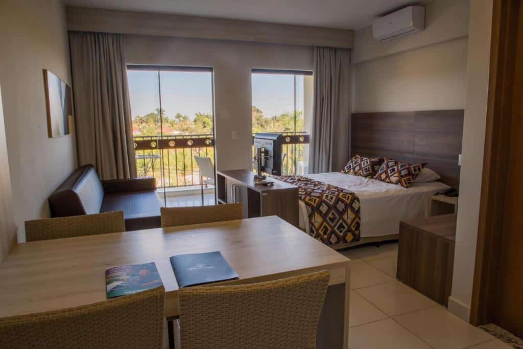 Quarto no Barretos Country Thermas Resort com uma cama de casal de frente para a tv, um sofá, uma mesa de madeira e sacada com portas de vidro, para representar hotéis em Barretos.