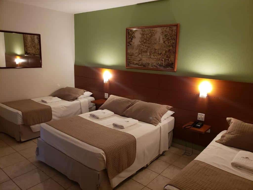Quarto no Proença Hotel com três camas, uma de casal e duas de solteiro, com um quadro na parede verde e um espelho na parede branca, para rperesentar hotéis em Barretos.
