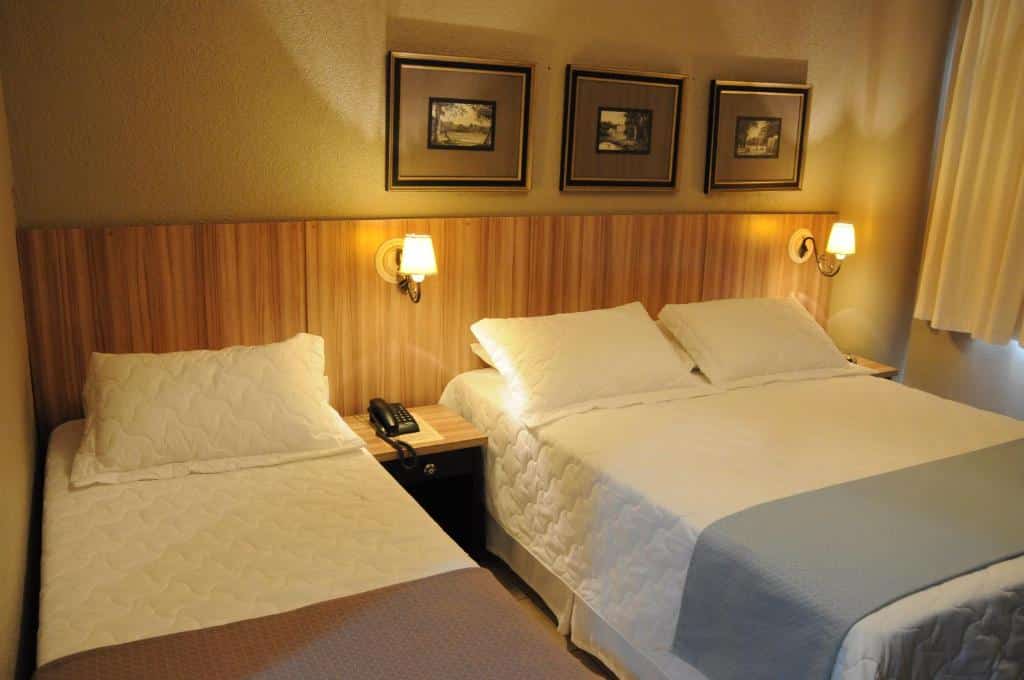 Quarto com luz amarela e duas camas, uma de casal e uma de solteiro com 3 quadros na parede e duas luminárias, no Hotel Kehdi Plaza, para representar hotéis em Barretos.