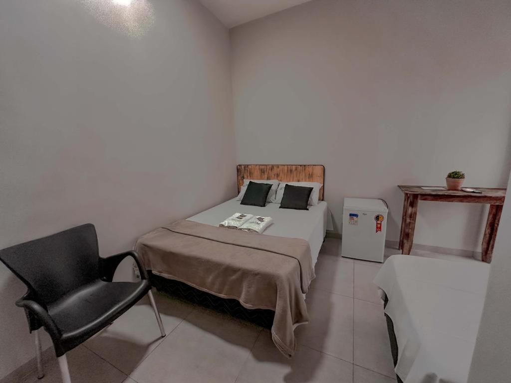 Um pequeno quarto com uma cama de casal, uma cadeira preta, um frigobar, uma mesa de madeira e um vaso de plantas, no Hotel Rosa Viva, para representar hotéis em Barretos.