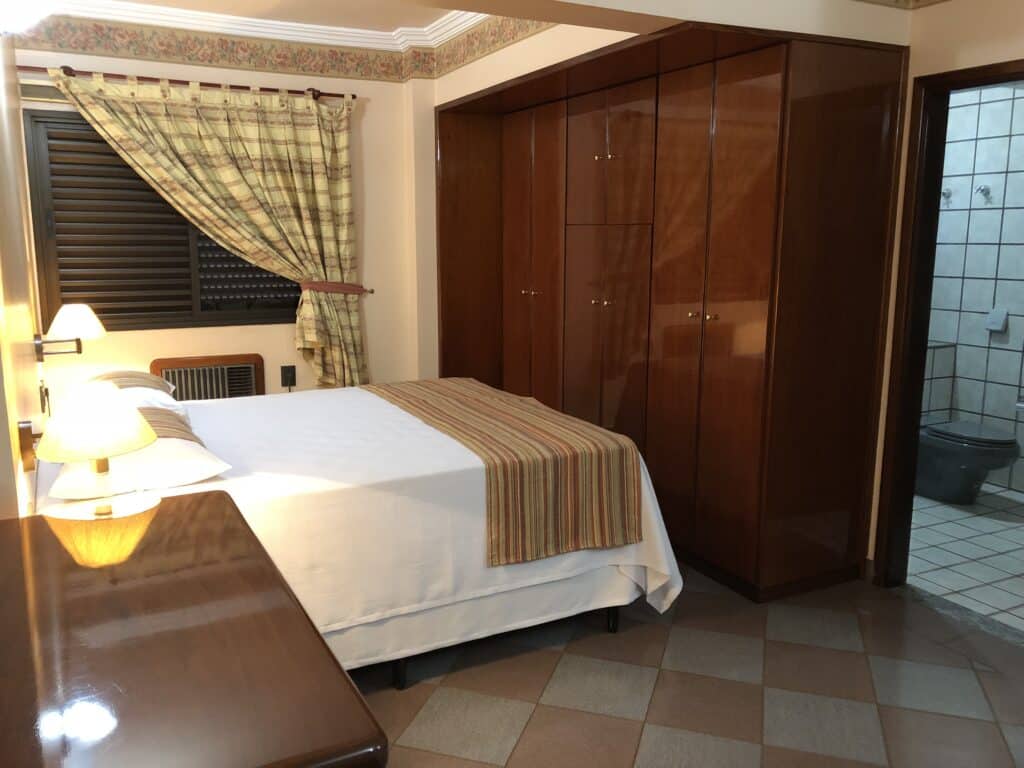 Quarto do Mabruk Barretos Apart Hotel mostrando uma cama de casal à esquerda da imagem, com um grande armário de madeira em frente aos pés da cama e uma porta mostrando o banheiro à direit, para representar hotéis em Barretos.