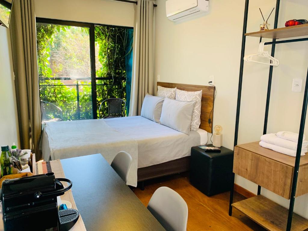 Um pequeno quarto com uma cama de casal, uma janela com vegetação externa, uma mesa, prateleiras abertas e um ar-condicionado, na Maritimus Suítes, para representar hotéis em Barretos.
