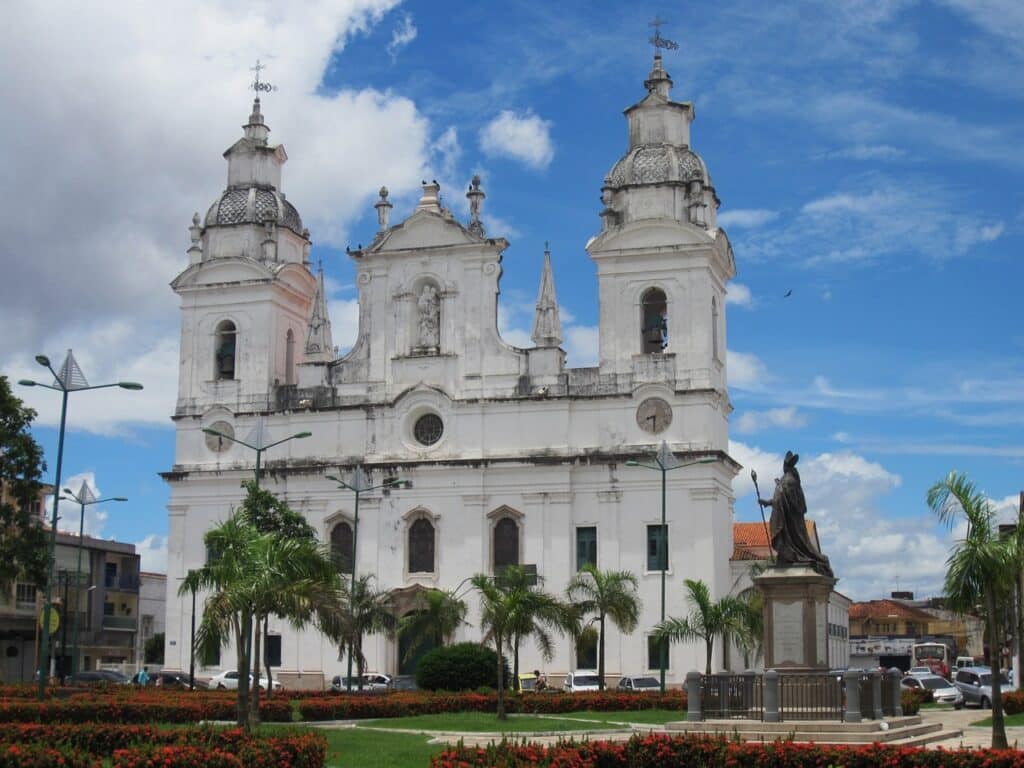 Catedral Metropolitana de Belém com um jardim, árvores e uma estátua logo a sua frente. Ao fundo é possível ver casas e carros. 