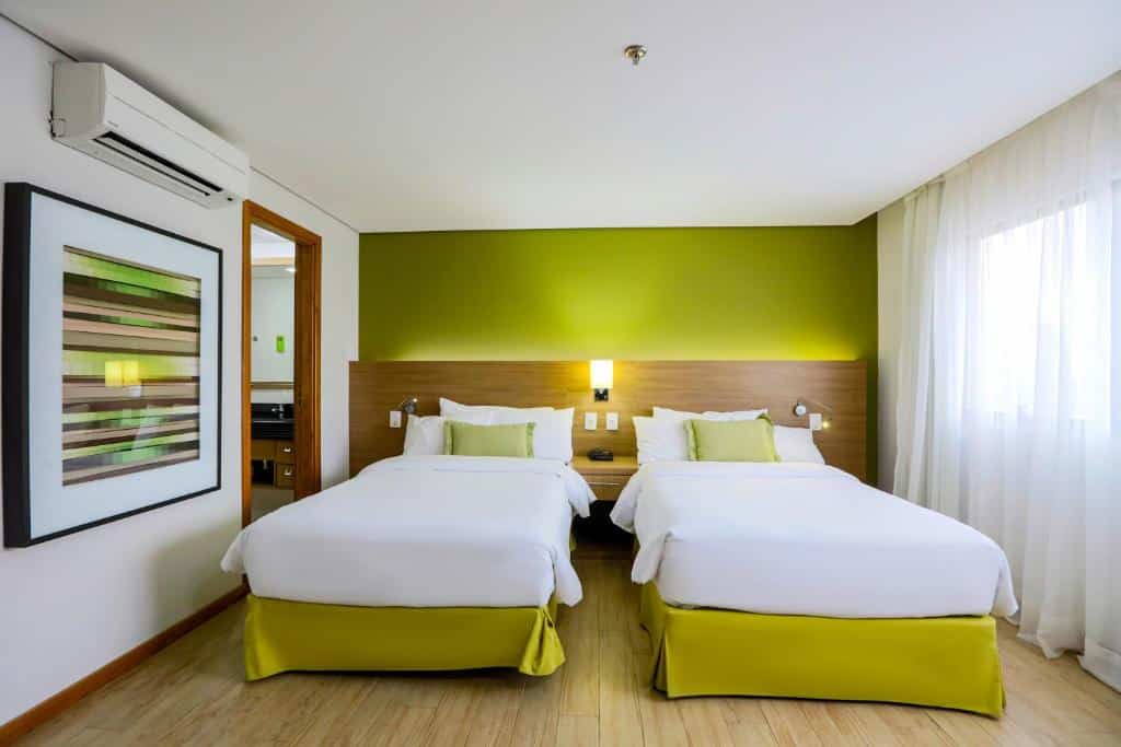 Quarto no Radisson Hotel Belém com duas camas, uma luminária no meio delas e janela com cortina do lado. No canto esquerdo há um ar condicionado, entrada para o banheiro e um quadro na parede, para representar hotéis em Belém do Pará.