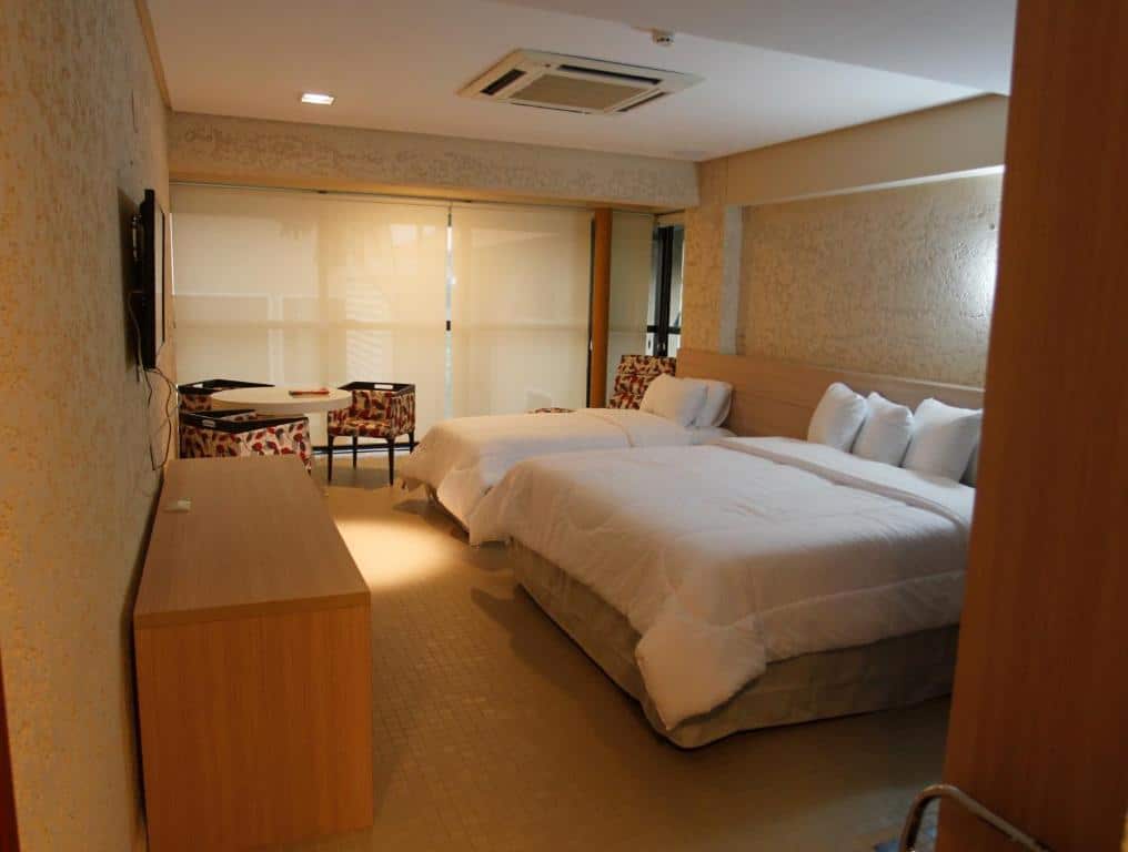 Quarto no Atrium Hotel Quinta de Pedras com duas camas, uma grande e outra de solteiro. Em frente a elas tem um móvel e uma televisão. Ao lado tem uma janela com cortinas brancas e uma mesinha com cadeiras, para representar hotéis em Belém do Pará.