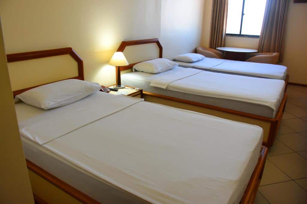 Quarto no Hotel Grão Para com três camas, um abajur em cima de uma mesinha de cabeceira com telefone. No canto há uma mesa redonda com duas cadeiras e uma janela com cortina, para representar hotéis em Belém do Pará.