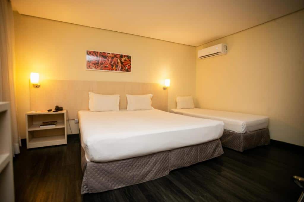 Quarto no ibis Styles Belem Batista Campos com duas camas, uma de casal e uma de solteiro. Há um quadro acima delas, duas luminárias e uma mesinha de cabeceira. É possível também ver um ar condicionado.