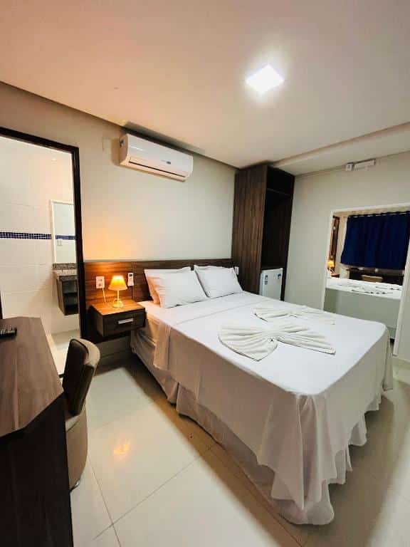 Quarto no Hotel Antiga Roma com uma cama, abajur, espelhos e mesa com cadeira. É possível ver um ar condicionado, uma cortina e, no canto, um mini frigobar, para representar hotéis em Belém do Pará.