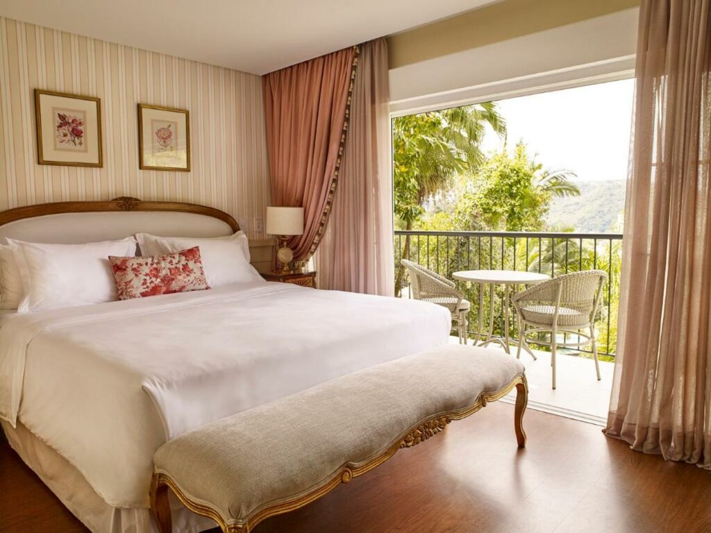 Um quarto no Villa do Vale Boutique Hotel com uma cama bem arrumada fica de frente para uma pequena varanda com duas cadeiras e uma mesa, com vista para a vegetação e as colinas do lado de fora, para representar hotéis em Blumenau.