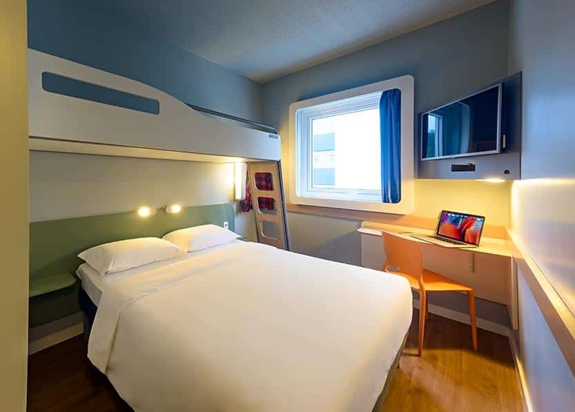 Um quarto compacto no Ibis Budget Blumenau com uma cama de casal, um beliche acima, uma escrivaninha com uma cadeira, uma TV e uma janela com cortinas azuis, para representar hotéis em Blumenau.