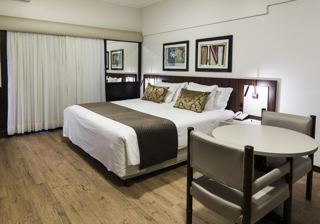 Quarto no Plaza Blumenau com uma cama grande, dois travesseiros, uma mesa redonda com cadeiras, piso de madeira e arte emoldurada na parede, para representar hotéis em Blumenau.