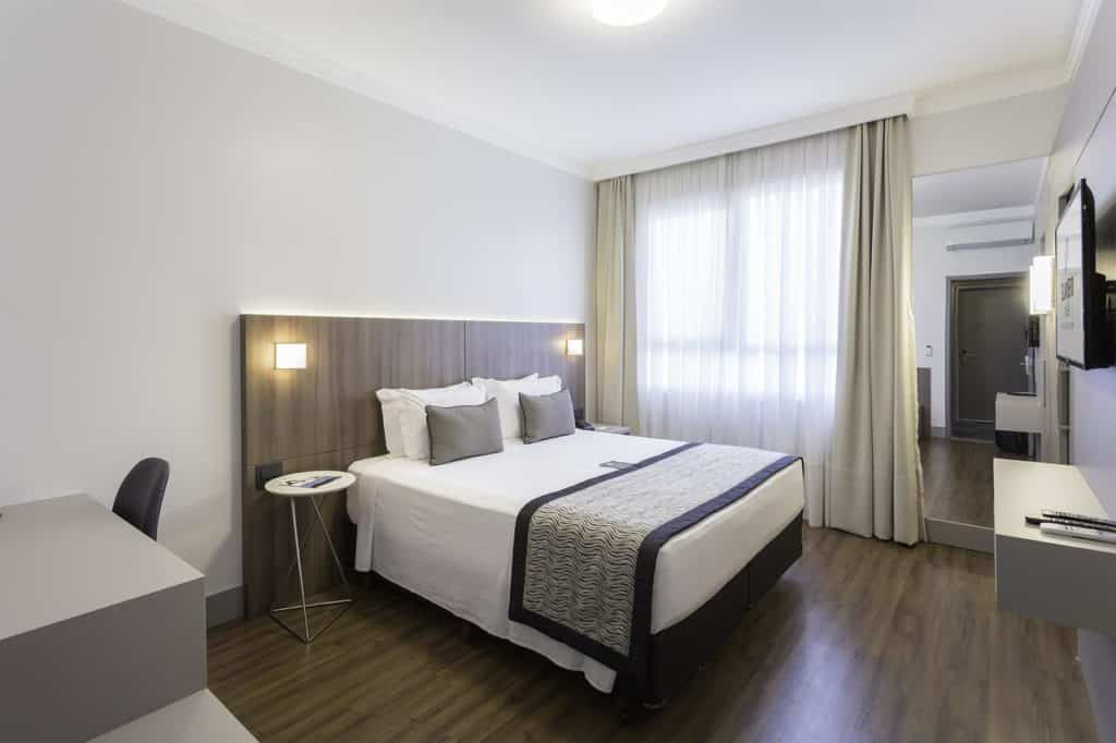 Quarto moderno no Slaviero Essential Blumenau com uma cama de casal, escrivaninha, TV, janela grande com cortinas e piso de madeira, para representar hotéis em Blumenau.