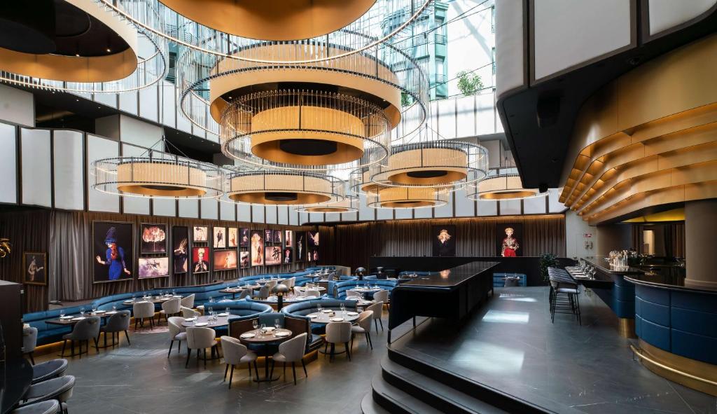 salão do Radisson Collection Grand Place Brussels, um dos hotéis em Bruxelas, com luminárias modernos enormes em formato redondo e tetos de vidro ao redor, com diversas mesas e cadeiras e um pavimento mais elevado com um balcão