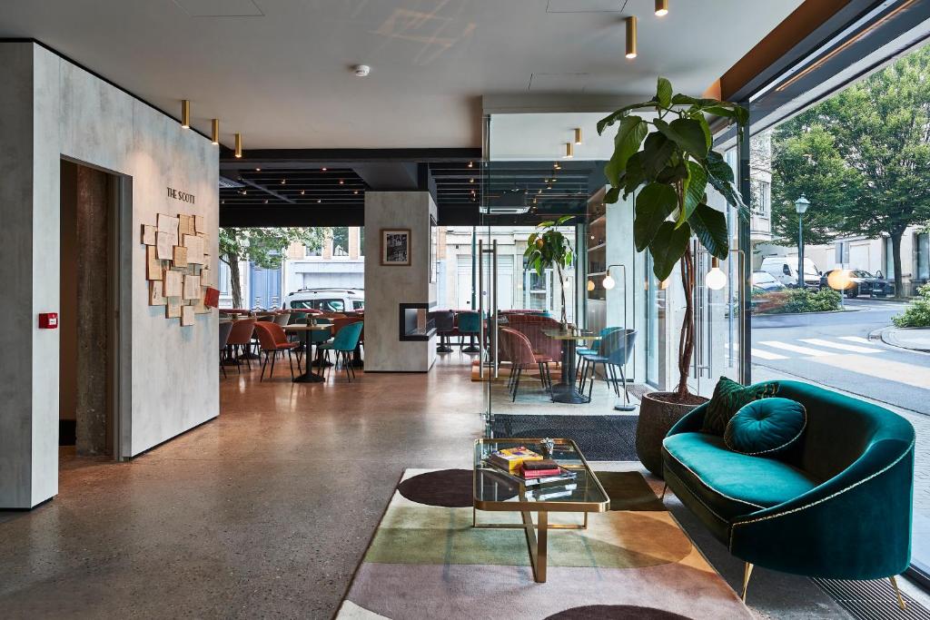 recepção com decoração sofisticada no The Scott Hotel Brussels, um os hotéis em Bruxelas, com porta ao lado de vários livros na parede, de um lado, há um sofá com mesas e, do outro, mesas e cadeiras para o buffet e portas de vidro que dão para a rua