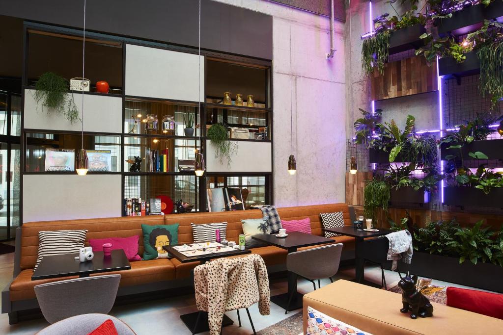 salão do Moxy Brussels City Center, um dos hotéis em Bruxelas, com luz arroxeada e sofá comprido com almofadas coloridas e com desenhos diferentes, com um jardim vertical que pega a parede inteiro do um lado e, mais ao centro, um bar também na vertical com outras cadeiras e mesas e luminárias