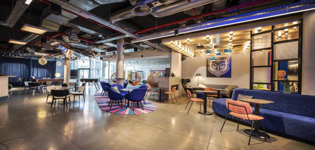 lounge do Aloft Brussels Schuman, um dos hotéis em Bruxelas, com espaços amplos, poltronas azuis e mesas com tapetes arco-íris abaixo de cada mesa, balcão atrás com quadros coloridos e iluminação amarelada