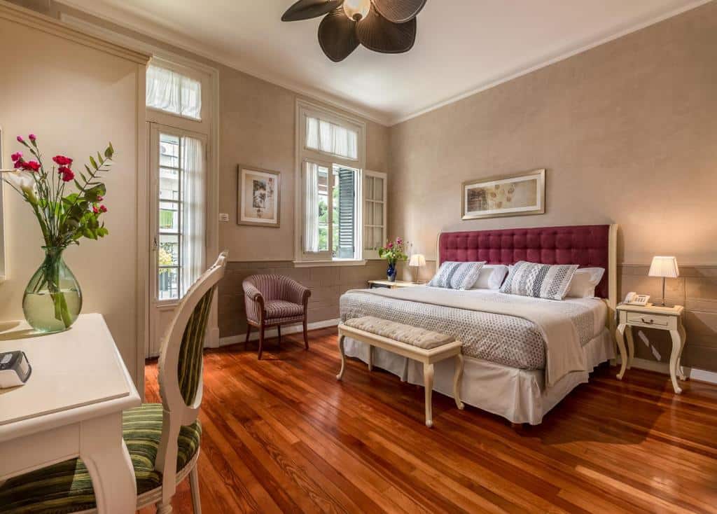 Quarto amplo no Duque Hotel Boutique & Spa com uma cama de casal, uma janela, uma porta que leva para a sacada, uma poltrona, duas mesinhas de cabeceira com abajures, uma mesa de escritório com uma cadeira, tudo em estilo de época, para representar hotéis em Buenos Aires