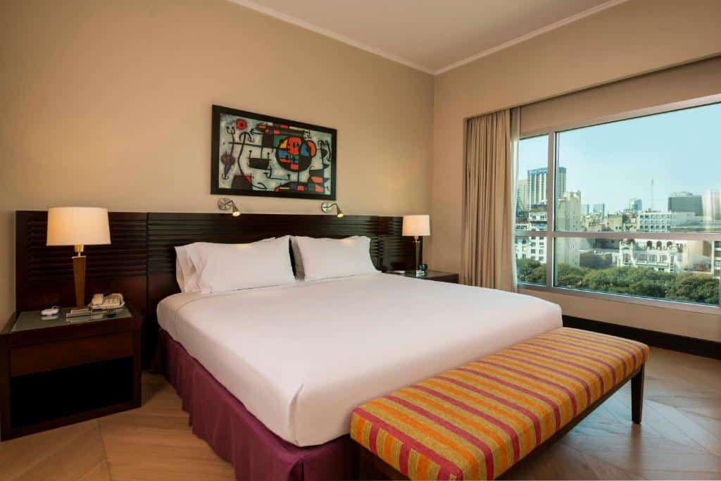 Quarto do Eurobuilding Hotel Boutique Buenos Aires com uma cama de casal, duas mesas de cabeceira com abajures, uma janela bem ampla com vista para a cidade com cortinas, o chão é de madeira, para representar os hotéis em Buenos AIres.