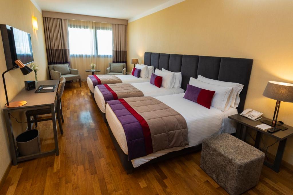 Quarto no Hotel Grand Brizo Buenos Aires com três camas de casal, uma escrivaninha, duas cadeiras ao lado de uma janela e piso de madeira, para representar hotéis em Buenos Aires.