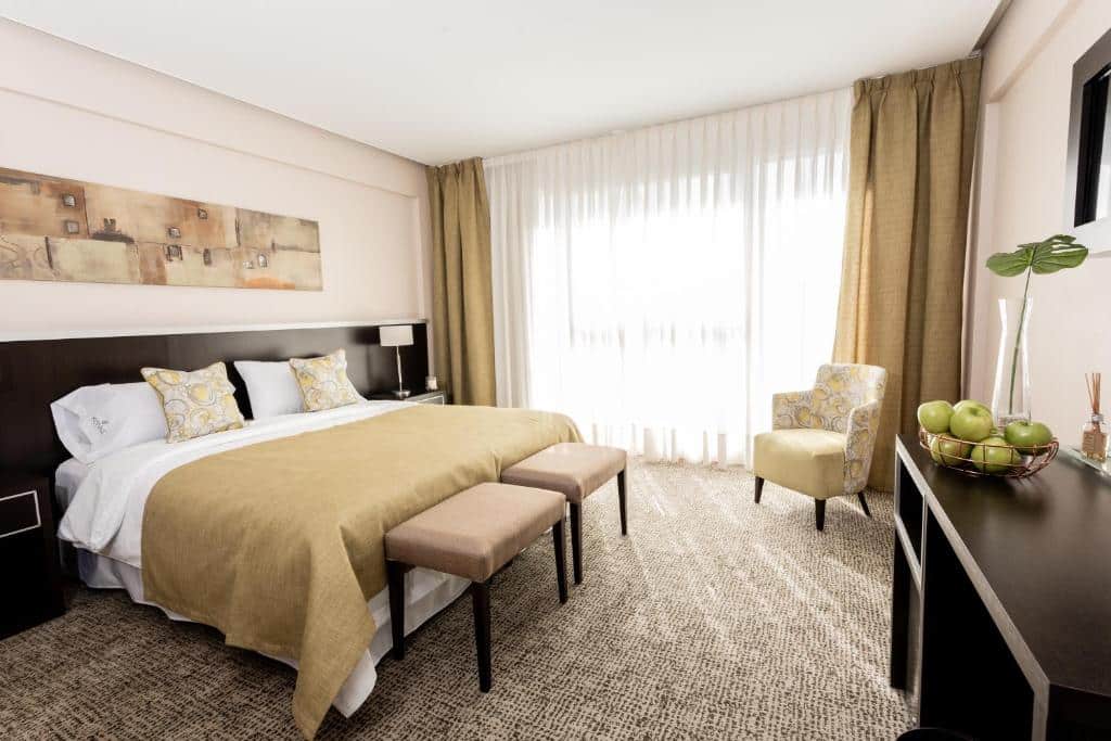 Quarto no Konke Buenos Aires Hotel com quarto amplo, uma cama de casal, uma sacada, uma poltrona, uma estante com um frigobar e algumas frutas em cima, o local é decorado em tons de bege, para representar hotéis em Buenos Aires.