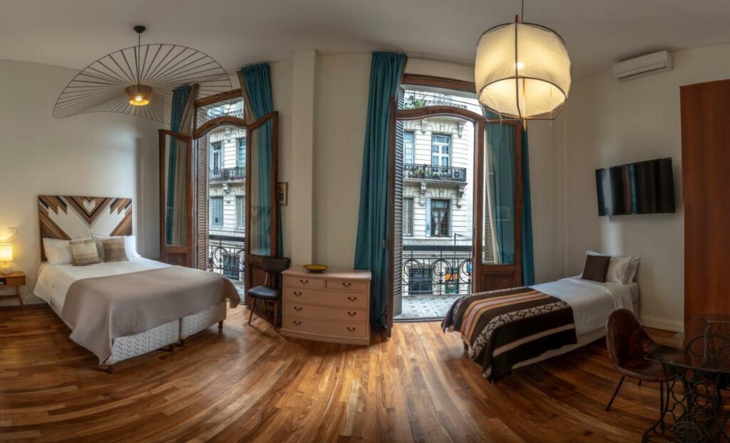 Quarto do L'Adresse Hôtel Boutique com uma cama de casal, uma de solteiro, duas sacadas com vista para a cidade, dois lustres artesanais, chão de madeira, uma cômoda, uma televisão, um pequeno armário, uma mesinha de escritório com uma cadeira e um ar-condicionado, tudo em tons de madeira e em estilo contemporâneo, para representar hotéis em Buenos AIres