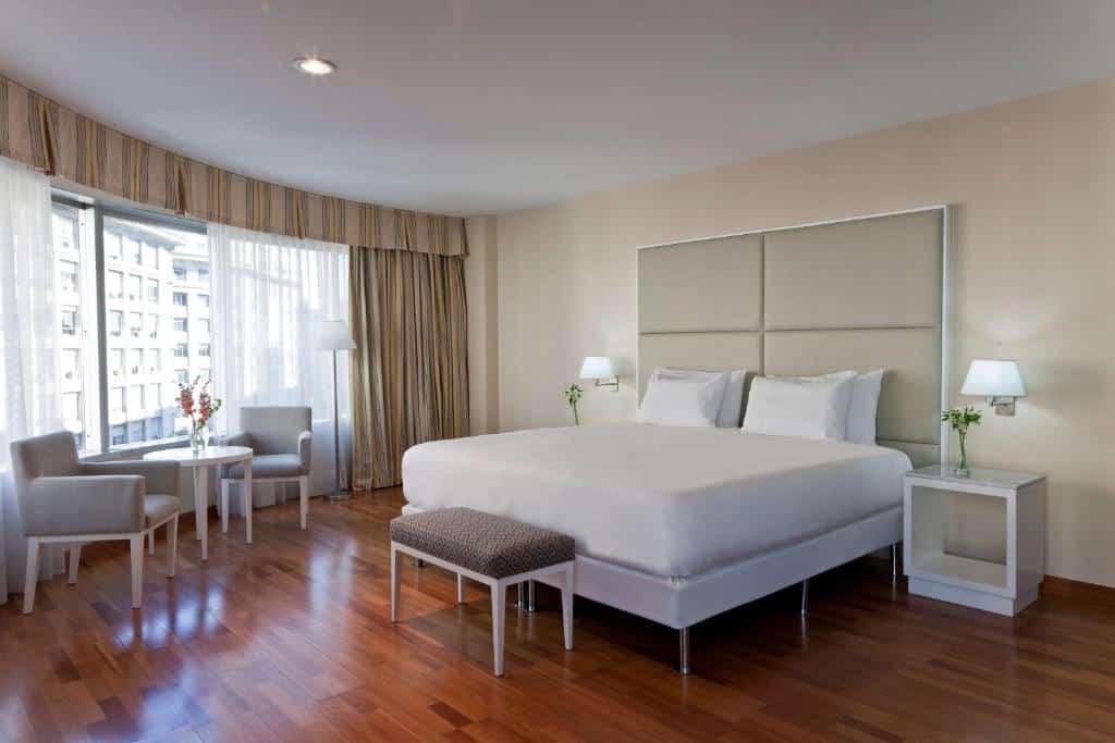 Quarto do NH Buenos Aires Latino com cama bem grande, mesinha e luminária de ambos os lados, em tons claros, janela grande que pega grande parte da parede e mostra bem a cidade, há uma mesa com cadeiras e o chão imita madeira, para rperesentar hotéis em Buenos Aires.