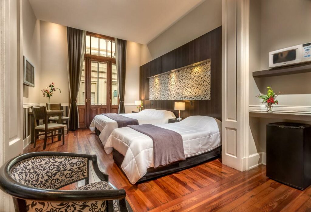 Quarto espaçoso do Up Tribeca, com uma cama de casal, uma de solteiro, piso de madeira, uma mesinha com duas cadeiras, uma poltrona, em uma parte separada do quarto há um frigobar e um micro-ondas