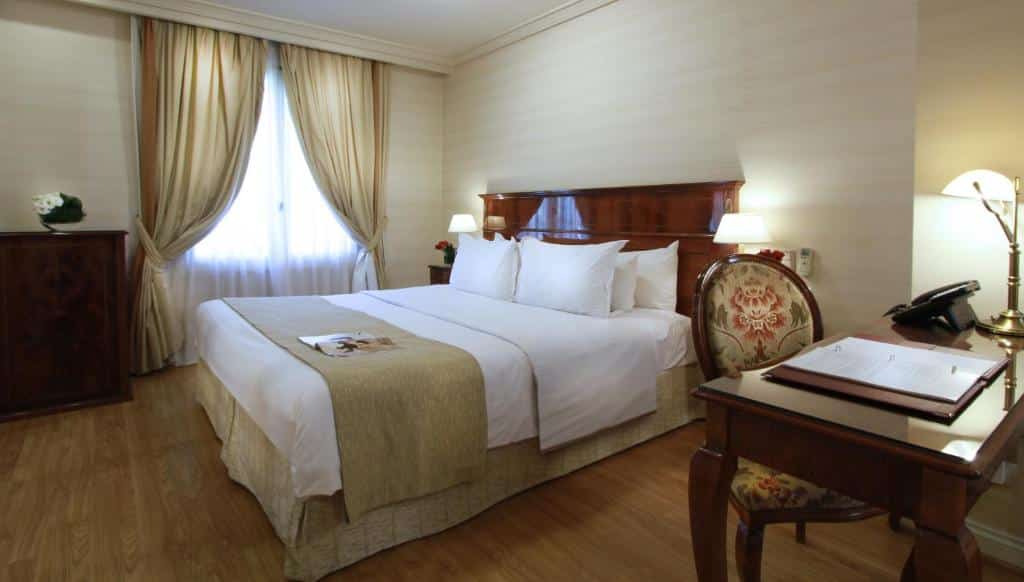 Quarto do Melia Recoleta Plaza Hotel com uma cama de casal, uma mesa de escritório com uma cadeira, uma janela com cortinas e uma cômoda, tudo em madeira e roupas de cama em bege e branco, para representar hotéis em Buenos Aires.