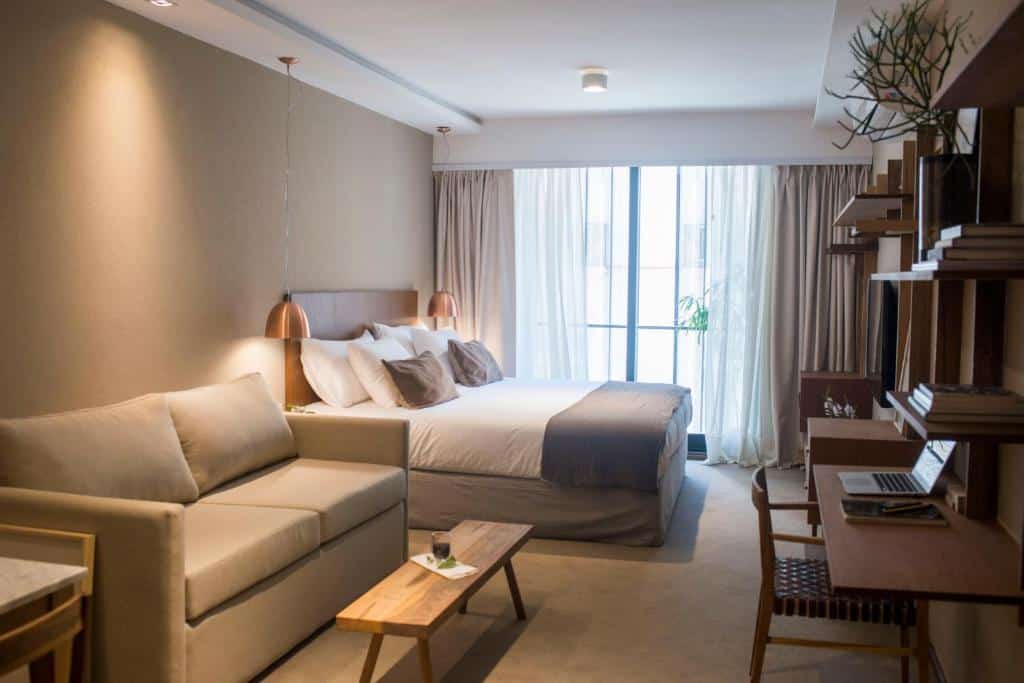 Suites da AQ Tailored Suitescom uma cama de casal, uma sacada, um sofá, uma mesinha de centro comprida em madeira, uma mesinha de escritório de madeira e uma cadeira, tudo em tons de bege e com móveis em madeira, para representar hotéis em Buenos Aires.