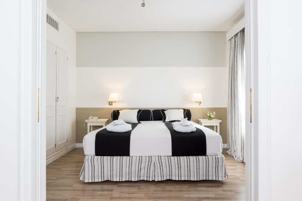 Quarto no Loi Suites Recoleta Hotel, com uma cama de casal centralizada, com travesseiros e toalhas sob a cama, duas mesinhas de cabeceira com vasos de flores, há duas luminárias presas na parede, do lado esquerdo, um armário embutido branco, o chão é de madeira.