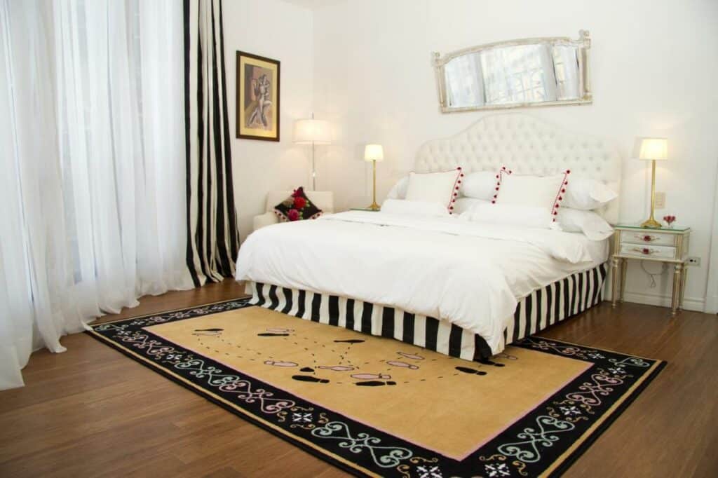 Quarto grande do Tango de Mayo Hotel com uma cama de casal, duas mesinhas de cabeceira com abajures, uma poltrona, uma janela com cortinas, chão de madeira e um tapete.