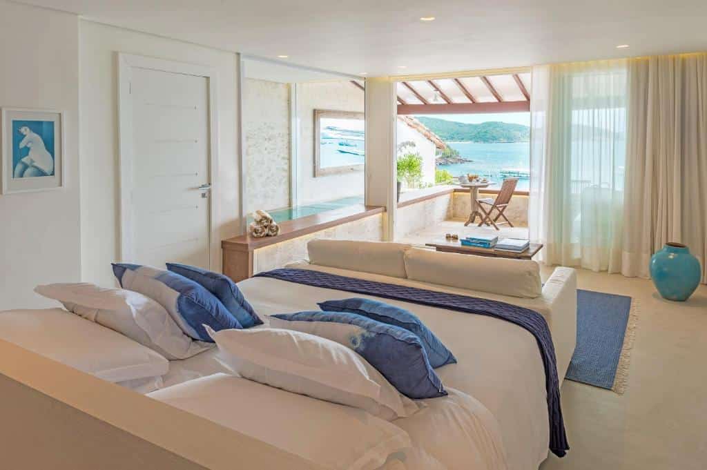 Quarto bem iluminado no Casas Brancas Boutique Hotel & Spa na Praia da Armação com uma cama grande, detalhes em azul, um sofá e uma vista aberta para uma varanda com vista para a água e as colinas, para representar hotéis em Búzios.