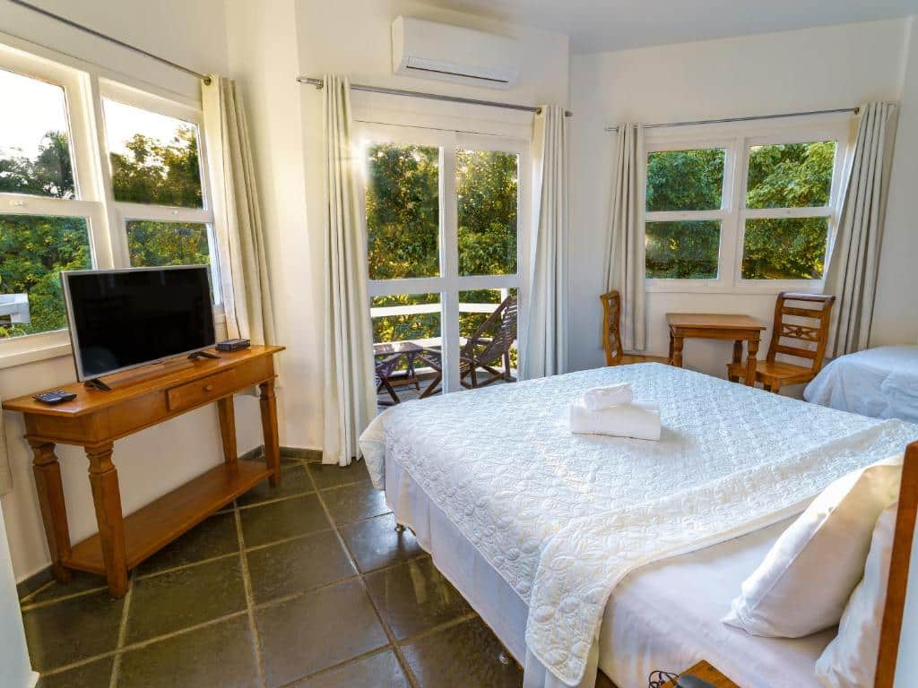 Um quarto bem iluminado na Apa Pau Brasil na Praia das Caravelas, com uma cama grande, uma TV em uma mesa de madeira, cadeiras e janelas grandes que mostram a vegetação do lado de fora, para representar hotéis em Búzios.