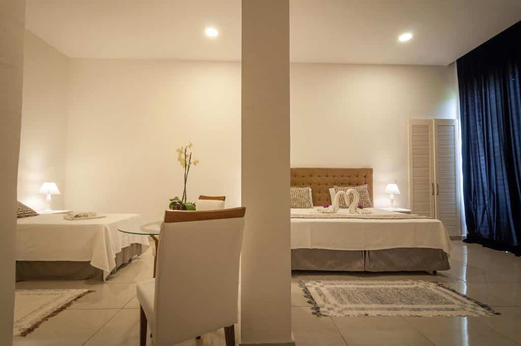Duas camas de casal em um quarto do Greco Hotel na Praia Brava com paredes brancas, piso de azulejo e móveis simples, separados por uma coluna central, para representar hotéis em Búzios.