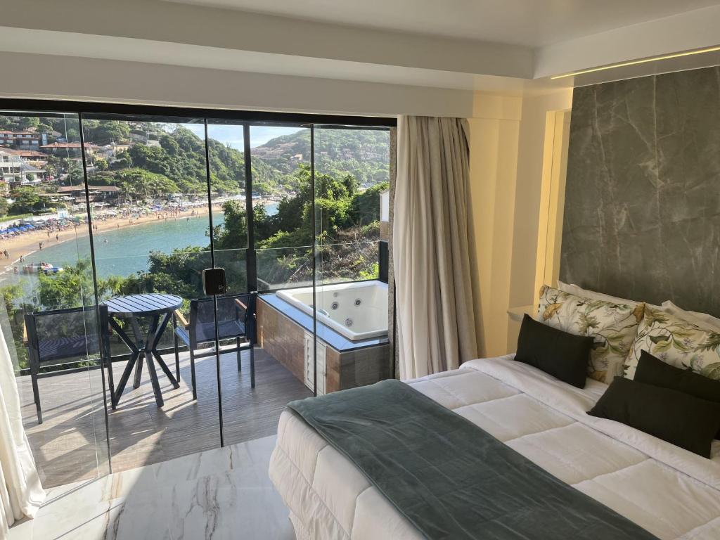 Quarto no Colonna Park Hotel na Praia João Fernandes com uma cama, uma pequena mesa e cadeiras na varanda, uma banheira de hidromassagem e vista para uma praia e colinas verdes do lado de fora, para representar hotéis em Búzios.