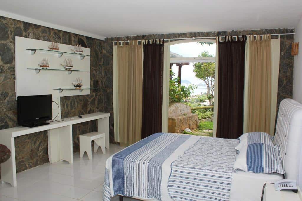 Um quarto no Bliss Hotéis Bella Vista na Praia Brava com colcha listrada, escrivaninha com TV, parede com detalhes em pedra e uma grande janela com vista para as árvores e a água, para representar hotéis em Búzios.