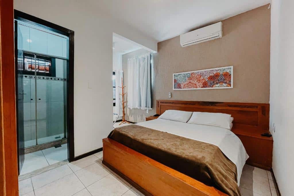 Um quarto no Búzios Centro Hotel no Centro, com uma cama de madeira, cobertor marrom, arte na parede, ar-condicionado e um banheiro privativo com chuveiro de vidro, para representar hotéis em Búzios.