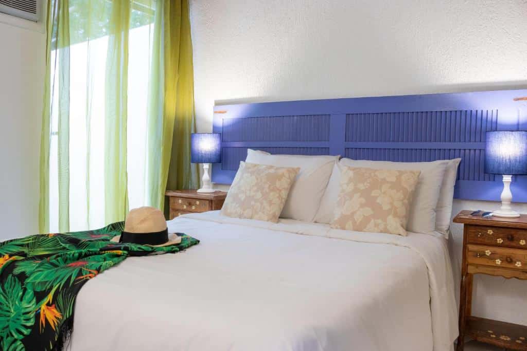 Uma cama bem arrumada com roupas de cama brancas, cabeceira azul, mesas de cabeceira e abajures; um chapéu e um xale sobre a cama, no Calador Búzios Hotel no Centro, para representar hotéis em Búzios.