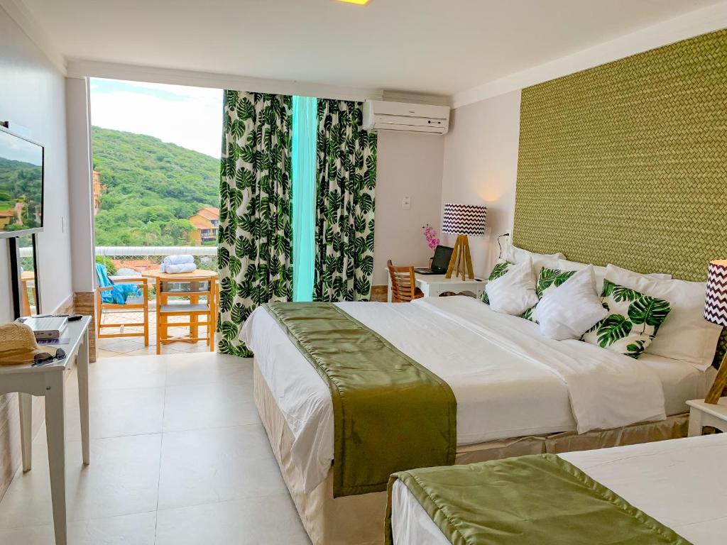 Um quarto no Hotel Pousada Experience João Fernandes na Praia João Fernandes com duas camas, detalhes em verde, uma TV, uma escrivaninha e uma varanda com vista para as colinas verdes, para representar hotéis em Búzios.