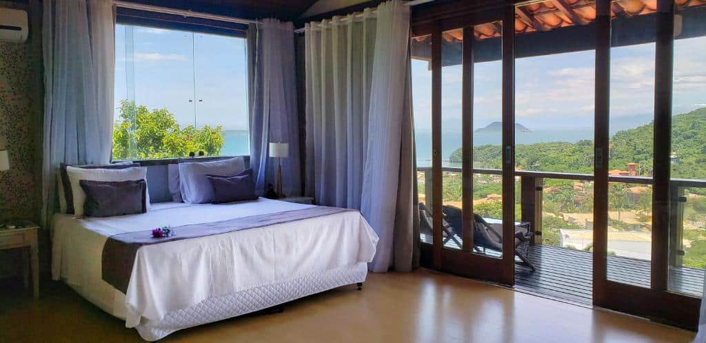 Um quarto na Ilha Branca Exclusive Hotel na Praia João Fernandes com uma cama de casal, janelas grandes, cortinas e uma varanda com vista para as colinas verdes e o oceano, para representar hotéis em Búzios.
