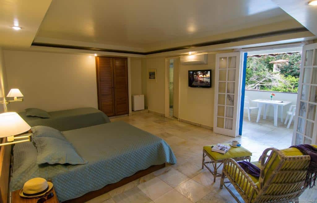 Quarto no Hotel Doce Mar na Praia da Armação com duas camas, uma cadeira, TV e portas abertas que dão para uma varanda com uma mesa e cadeiras brancas, para representar hotéis em Búzios.