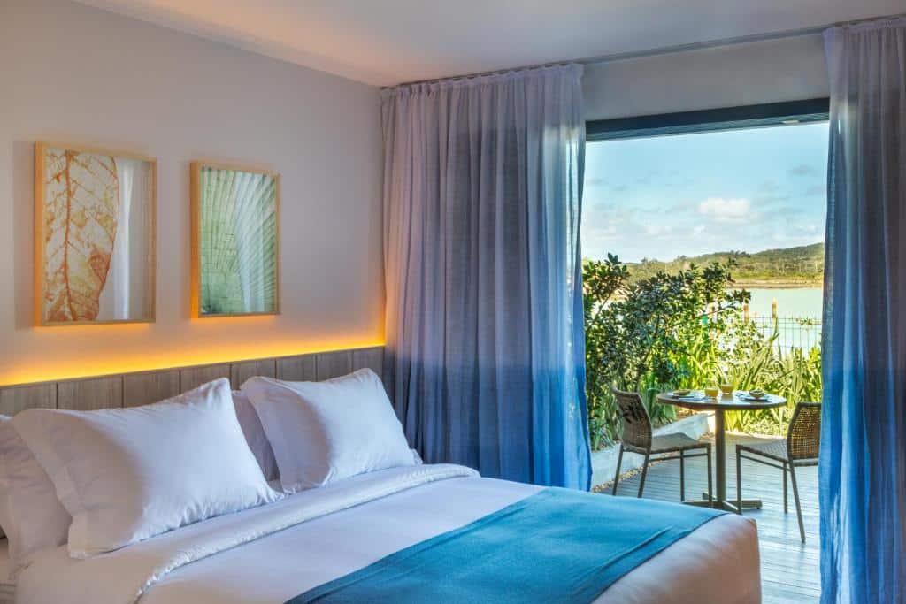 Um quarto do Hotel Aretê na Praia Rasa com uma cama bem arrumada, duas obras de arte na parede e uma varanda com assentos ao ar livre com vista para a vegetação e a água, para representar hotéis em Búzios.
