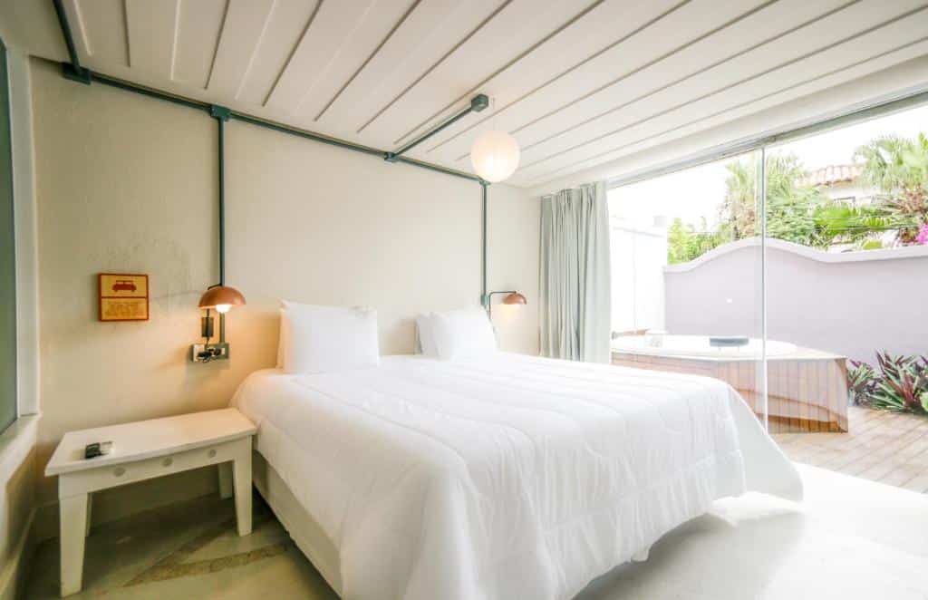 Quarto minimalista no Selina Buzios na Rua das Pedras com uma cama branca, mesa lateral, luminárias de parede e uma grande janela com vista para um pátio com banheira de hidromassagem, para representar hotéis em Búzios.