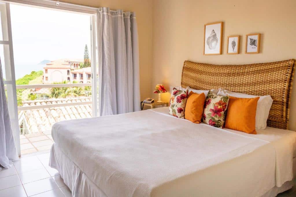 Um quarto bem iluminado no Bahiamarela Boutique Hotel & SPA na Praia João Fernandes, com uma cama grande, cabeceira de vime, travesseiros coloridos e uma janela aberta com vista para árvores e prédios, para representar hotéis em Búzios.