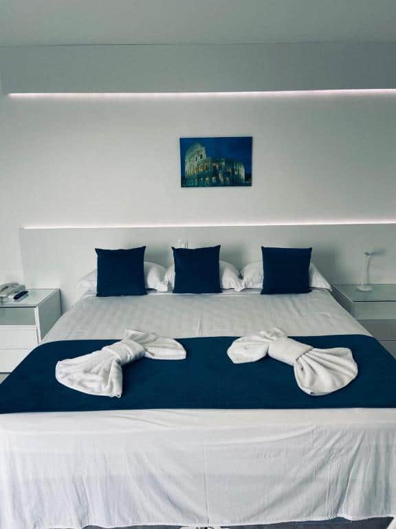 Quarto no Hotel Real com cama de casal com travesserios, almofadas, toalhas, uma fita de led na cabeçeira da cama e um quadro decorativo acima, para representar hotéis em Cabo Frio.