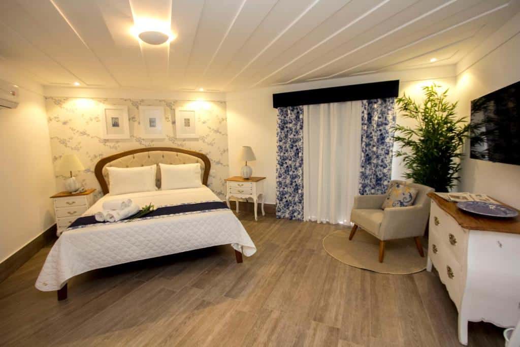 Grande quarto no Hotel Boutique Recanto da Passagem com cama de casal, decoração em tons de azul e branco, abajour, poltrona, mesinhas, cômoda e televisão, para representar hotéis em Cabo Frio.