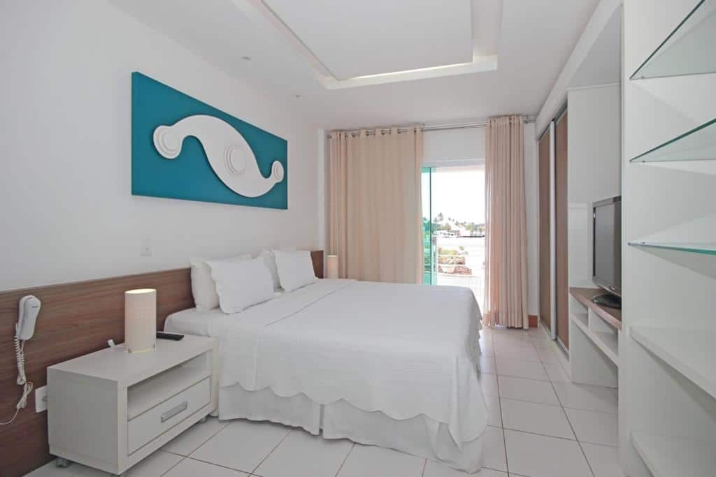 Quarto do Hotel Paradiso del Sol com cama de casal, cortinas beje, móveis brancos, televisão, quadro azul e varanda, para representar hotéis em Cabo Frio.