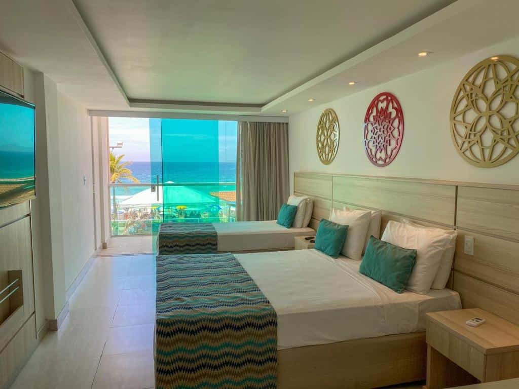 Quarto no Paradiso Peró Praia Hotel com uma casa de casal, uma casa de solteiro, cortinas beje, móveis em tons de marrom claro e varanda com vista para o mar, para representar hotéis em Cabo Frio.