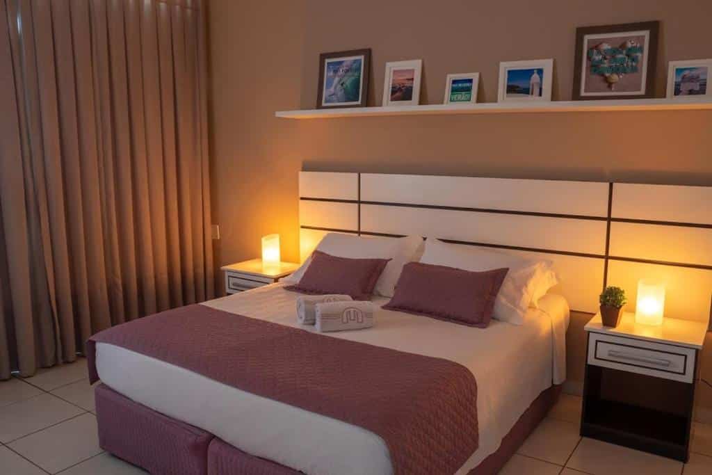 Quarto no Hotel Marlen com cama de casal, cabeçeira e mesinhas brancas, prateleira com quadros decorativos e cortina, para representar hotéis em Cabo Frio.